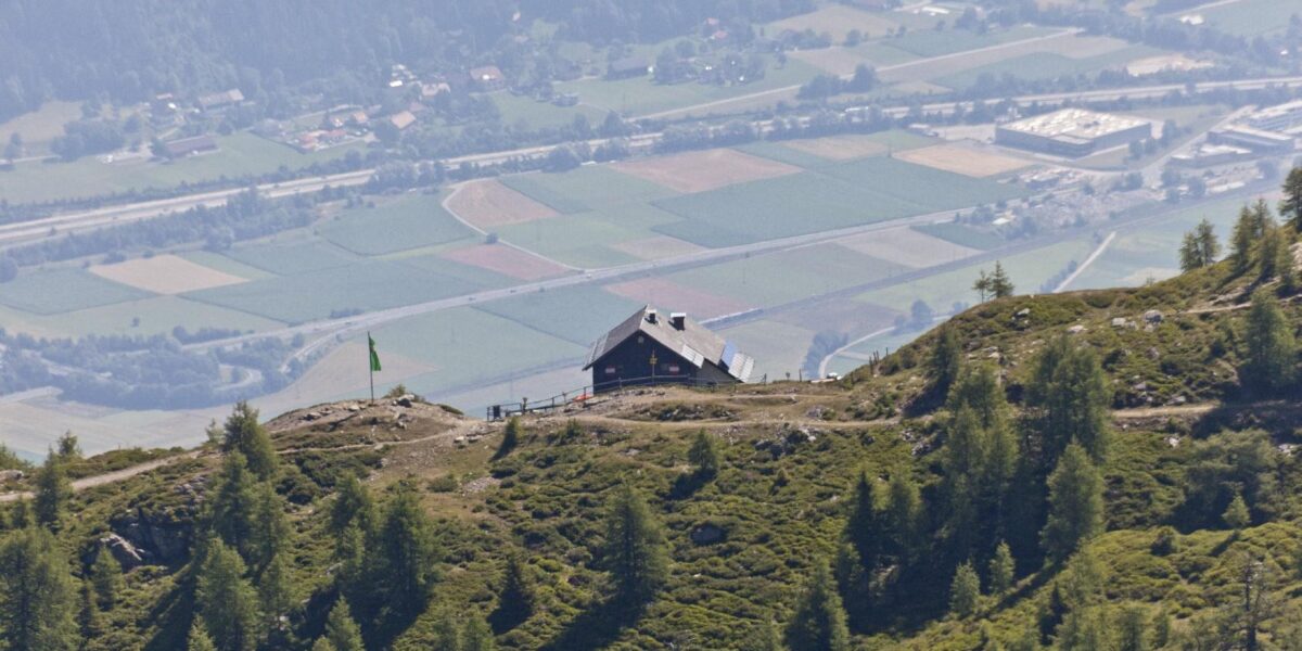 Eine Berghütte liegt auf einem grünen Hügel mit Blick auf ein Tal, in dem im Hintergrund Felder, Bäume und Gebäude zu sehen sind.