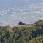 Eine Berghütte liegt auf einem grünen Hügel mit Blick auf ein Tal, in dem im Hintergrund Felder, Bäume und Gebäude zu sehen sind.