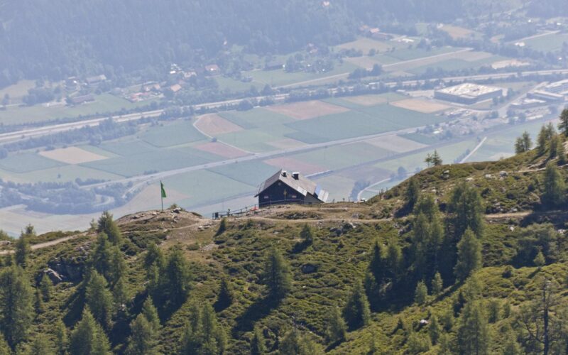 Eine Berghütte liegt auf einem grünen Hügel mit Blick auf ein Tal, in dem im Hintergrund Felder, Bäume und Gebäude zu sehen sind.