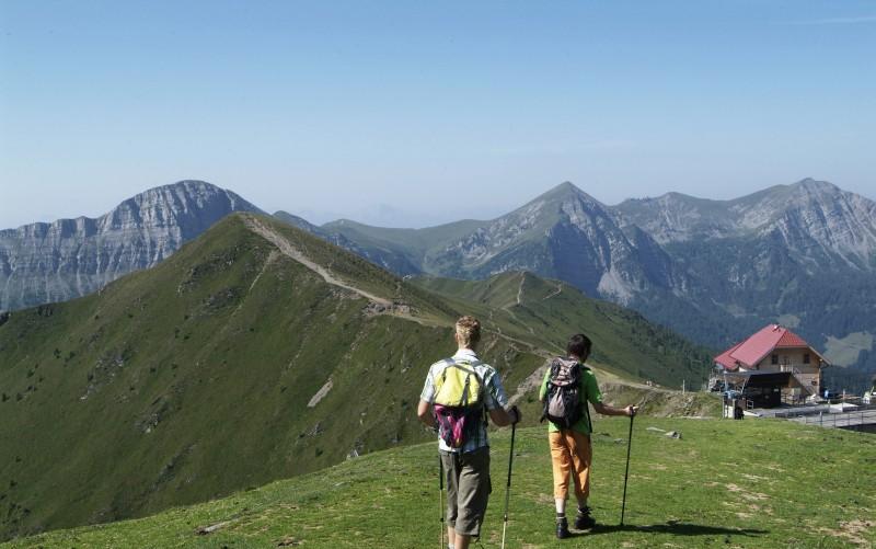 Zwei Wanderer mit Rucksäcken und Wanderstöcken gehen entlang eines grasbewachsenen Bergrückens mit Gipfeln und einem Gebäude im Hintergrund unter einem klaren Himmel.
