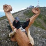 Zwei Personen sitzen auf einer großen, geschnitzten Holzbank auf einem felsigen Hügel, im Hintergrund sind Gebäude und eine hohe Antenne zu sehen.