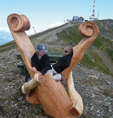 Zwei Personen sitzen auf einer großen, geschnitzten Holzbank auf einem felsigen Hügel, im Hintergrund sind Gebäude und eine hohe Antenne zu sehen.