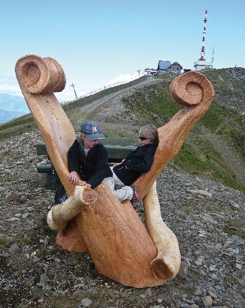 Zwei Personen sitzen auf einer großen, geschnitzten Holzbank auf einem felsigen Hügel, im Hintergrund sind Gebäude und eine hohe Antenne zu sehen.