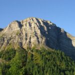 Felsengebirge mit ausgeprägten Schichtungen unter einem klaren blauen Himmel, mit grünen Kiefern an den unteren Hängen.