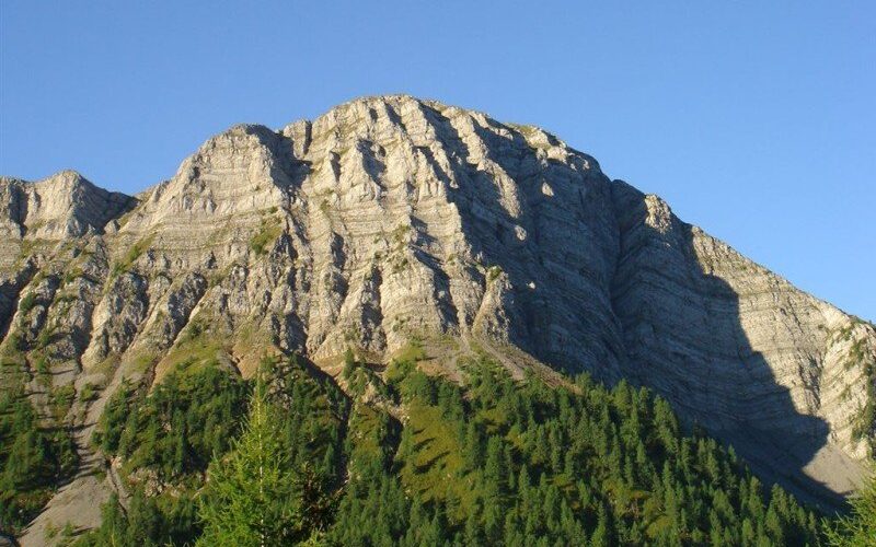 Felsengebirge mit ausgeprägten Schichtungen unter einem klaren blauen Himmel, mit grünen Kiefern an den unteren Hängen.