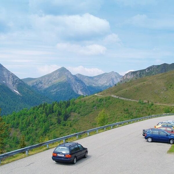 Mehrere Autos sind auf einem kleinen Parkplatz neben einer Bergstraße geparkt, mit grünen Hügeln und Gipfeln im Hintergrund und einem teilweise bewölkten Himmel.