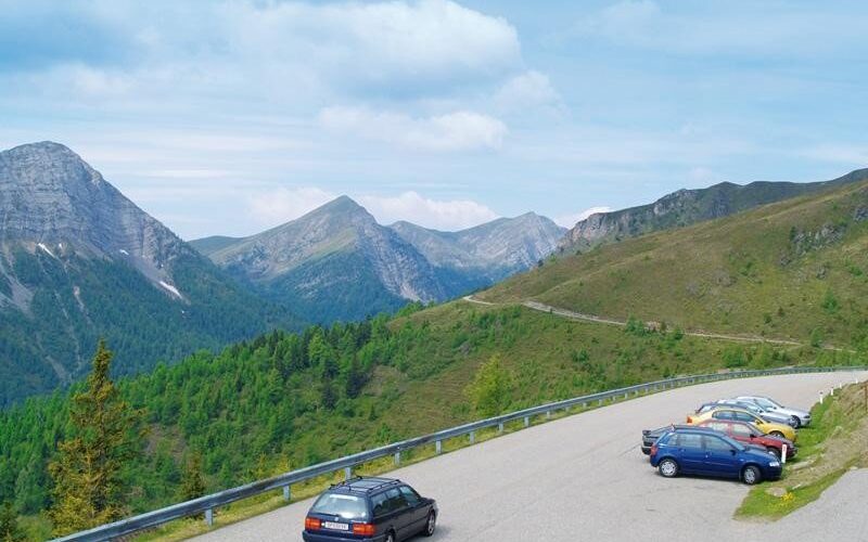 Mehrere Autos sind auf einem kleinen Parkplatz neben einer Bergstraße geparkt, mit grünen Hügeln und Gipfeln im Hintergrund und einem teilweise bewölkten Himmel.