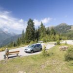 Ein kleiner Parkplatz mit einem Auto, Bänken und einer sitzenden Person, umgeben von Bäumen und Bergen unter einem klaren blauen Himmel.