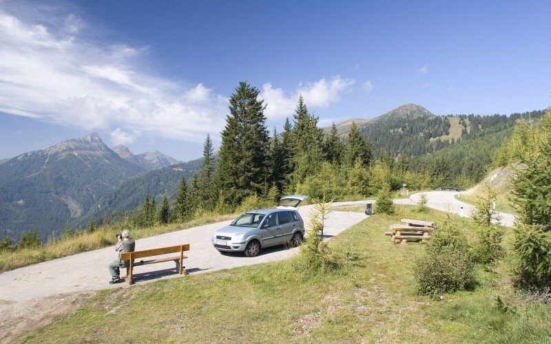 Ein kleiner Parkplatz mit einem Auto, Bänken und einer sitzenden Person, umgeben von Bäumen und Bergen unter einem klaren blauen Himmel.