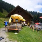 Holzhütte mit Sitzgelegenheiten im Freien, Sonnenschirmen und Spielzeug; Menschen sitzen und stehen in der Nähe der Bänke; grasbewachsener Hügel und Bäume im Hintergrund; österreichische Flagge sichtbar.