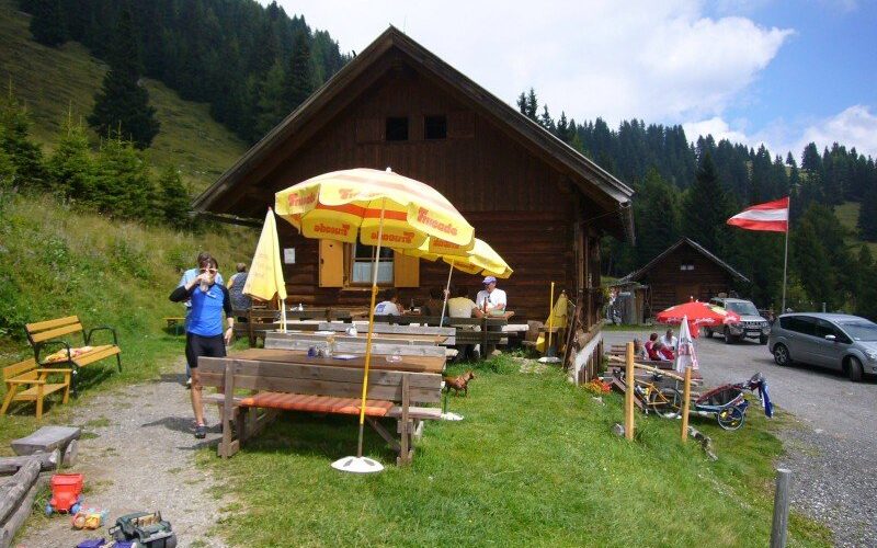 Holzhütte mit Sitzgelegenheiten im Freien, Sonnenschirmen und Spielzeug; Menschen sitzen und stehen in der Nähe der Bänke; grasbewachsener Hügel und Bäume im Hintergrund; österreichische Flagge sichtbar.