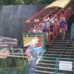 Die Menschen sitzen in einer Standseilbahn im Freien neben einer Treppe, mit Schildern und einer Cowboyfigur an der unteren Station.