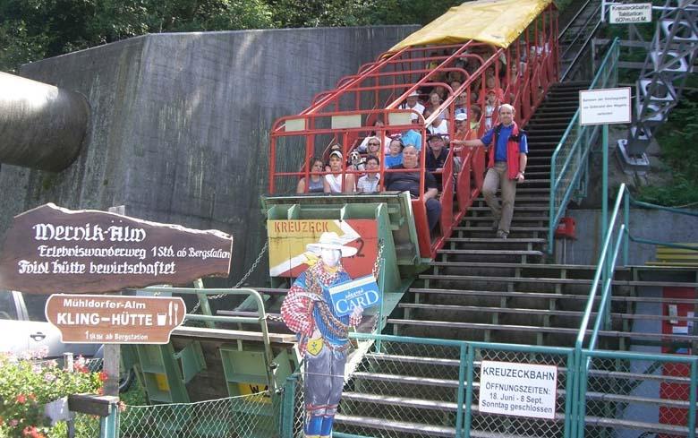 Die Menschen sitzen in einer Standseilbahn im Freien neben einer Treppe, mit Schildern und einer Cowboyfigur an der unteren Station.