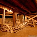 Ein hölzerner Pflug und andere alte landwirtschaftliche Geräte sind in einer schwach beleuchteten, rustikalen Holzscheune mit freiliegenden Balken und Säulen untergebracht.