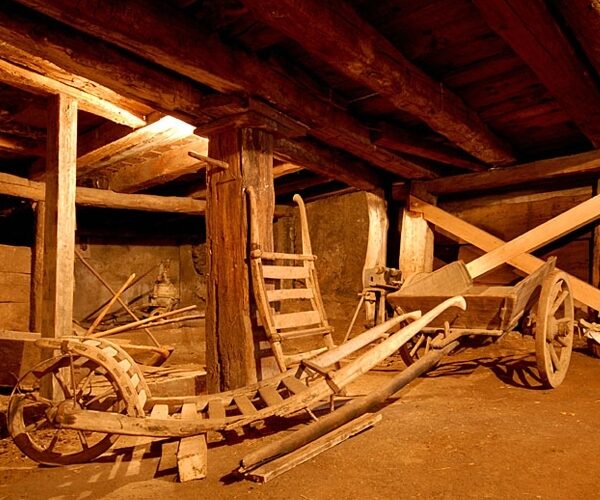 Ein hölzerner Pflug und andere alte landwirtschaftliche Geräte sind in einer schwach beleuchteten, rustikalen Holzscheune mit freiliegenden Balken und Säulen untergebracht.