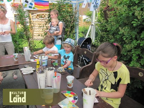 Die Kinder sitzen an einem Tisch im Freien und malen und basteln. Auf dem Tisch stehen Malutensilien und Getränke. Im Hintergrund stehen Erwachsene und ein Baby. Logo "Kultur am Land" in der unteren linken Ecke.