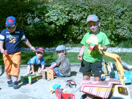 Vier kleine Kinder spielen mit Spielzeuglastwagen und Baufahrzeugen in einem Sandkasten im Freien, umgeben von Grün und Sonnenlicht.
