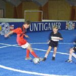 Drei Jungen spielen Fußball auf einem blauen Hallenfeld; einer in einem roten Trikot schießt den Ball, während zwei andere in schwarzen Trikots versuchen, ihn zu verteidigen.