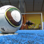 Nahaufnahme eines Fußballs auf einem blauen Hallenfeld, mit einem unscharfen Torwart, der auf das Tor im Hintergrund zuläuft.