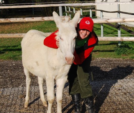 Eine Person mit rotem Hut und roter Jacke umarmt an einem sonnigen Tag einen weißen Esel im Freien in der Nähe eines Holzzauns.