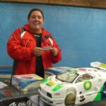 Eine Person in roter Jacke steht hinter einem Tisch mit ferngesteuerten Autos und einem Modellauto mit BP-Aufdruck, lächelt und hält einen Zettel in der Hand.