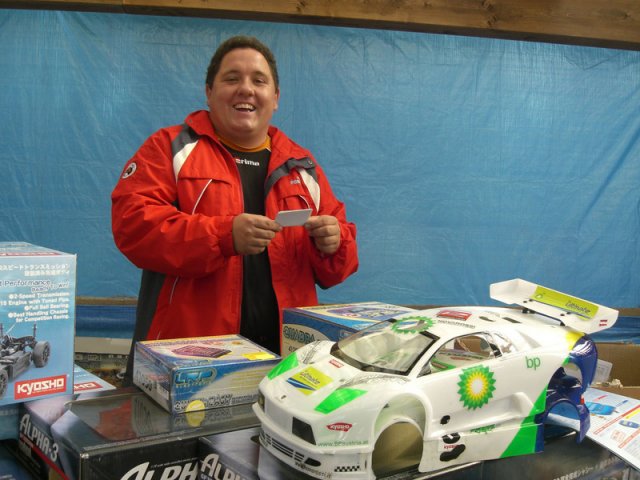 Eine Person in roter Jacke steht hinter einem Tisch mit ferngesteuerten Autos und einem Modellauto mit BP-Aufdruck, lächelt und hält einen Zettel in der Hand.