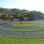 Eine Menschenmenge beobachtet an einem sonnigen Tag eine kleine, kurvenreiche Rennstrecke im Freien, umgeben von Gras und Bäumen.