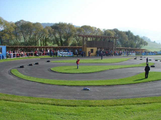 Eine Menschenmenge beobachtet an einem sonnigen Tag eine kleine, kurvenreiche Rennstrecke im Freien, umgeben von Gras und Bäumen.