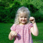 Ein junges Mädchen mit blonden Haaren und einem rotkarierten Hemd steht im Gras und hält einen kleinen Gegenstand in der Hand, während im Hintergrund Bäume und Büsche zu sehen sind.