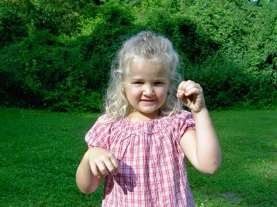 Ein junges Mädchen mit blonden Haaren und einem rotkarierten Hemd steht im Gras und hält einen kleinen Gegenstand in der Hand, während im Hintergrund Bäume und Büsche zu sehen sind.