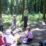 Eine Gruppe von Kindern sitzt auf Matten in einer Waldlichtung und spielt, während ein Erwachsener in der Nähe steht und sie beobachtet. Das Sonnenlicht fällt durch die Bäume.