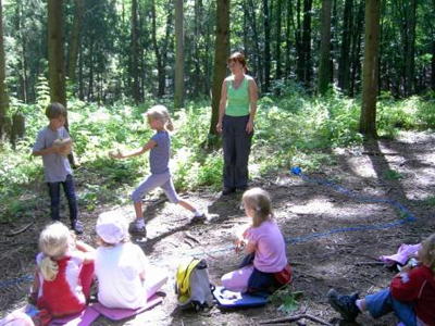 Eine Gruppe von Kindern sitzt auf Matten in einer Waldlichtung und spielt, während ein Erwachsener in der Nähe steht und sie beobachtet. Das Sonnenlicht fällt durch die Bäume.