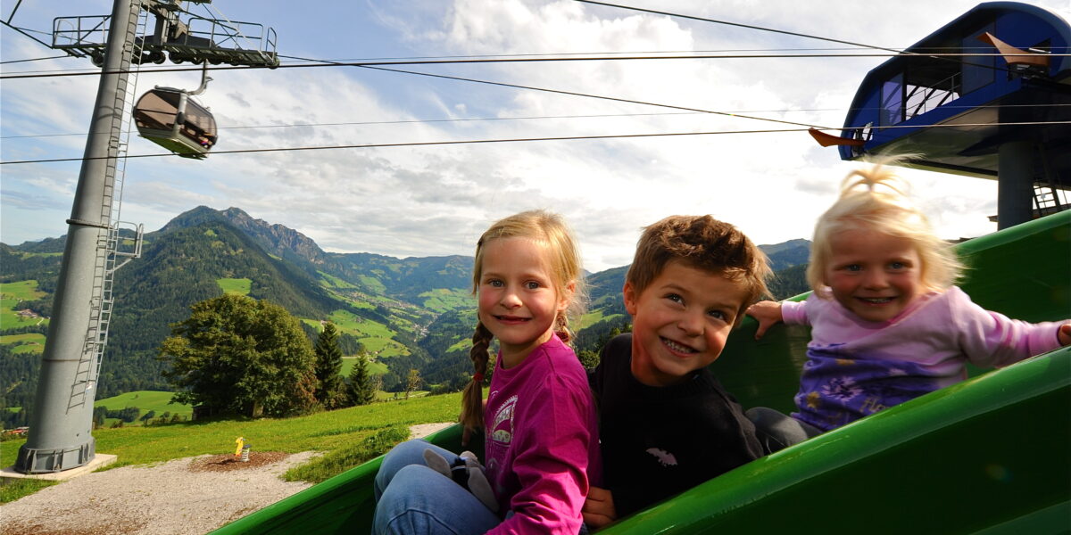 Drei kleine Kinder sitzen auf einer grünen Rutsche im Freien und lächeln in die Kamera. Hinter ihnen sind Berge, eine Gondelbahn und ein teilweise bewölkter Himmel zu sehen.