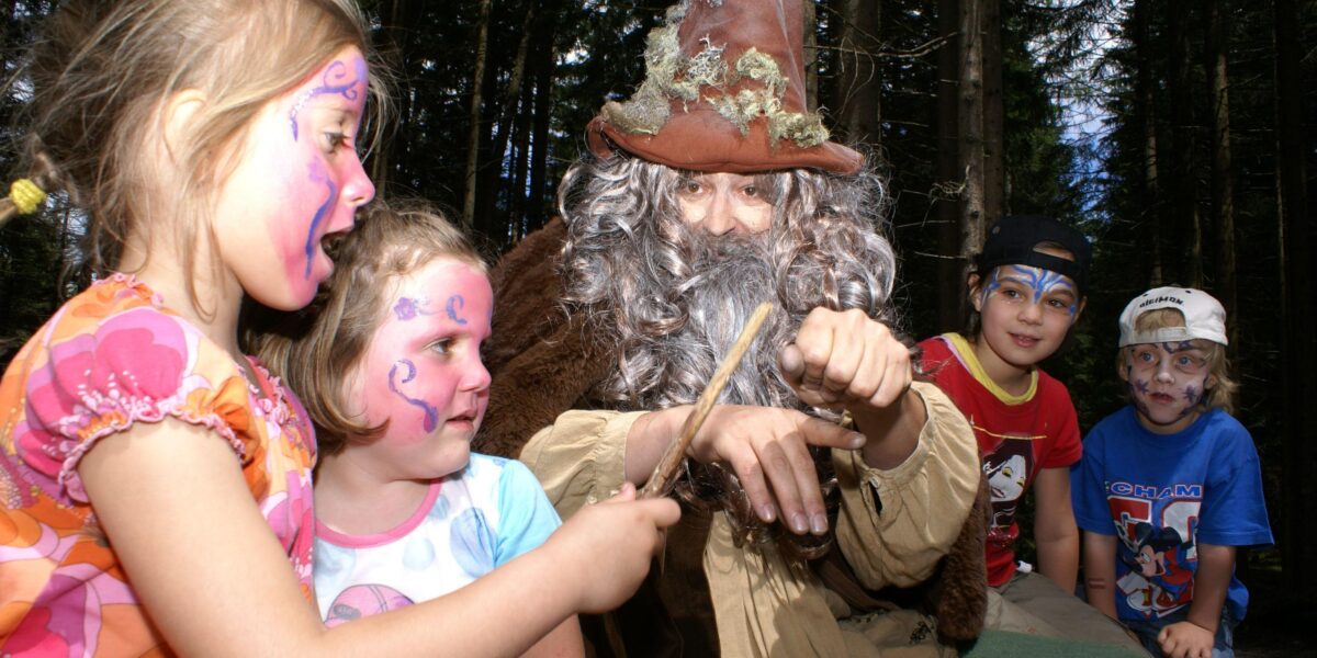 Kinder mit geschminkten Gesichtern interagieren mit einer als Zauberer verkleideten Person in einem Wald, die Stöcke hält und lächelt.