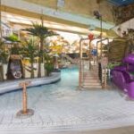 Indoor-Wasserpark mit einem flachen Becken, Palmen, Spielgeräten, einer Seilbrücke und einer lila Rutsche.