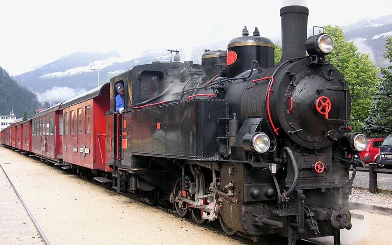 Eine schwarze Dampflokomotive mit roten Personenwagen hält an einem bewölkten Tag an einem Bahnhof. Im Führerstand des Zuges ist eine Person zu sehen.