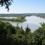 Blick auf einen breiten Fluss, der sich durch eine dicht bewaldete Landschaft schlängelt, unter einem klaren blauen Himmel, von einem erhöhten Aussichtspunkt aus gesehen.