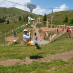 Ein Kind fährt auf einer Seilrutsche auf einem Spielplatz im Freien mit grasbewachsenen Hügeln und mehreren Personen im Hintergrund.