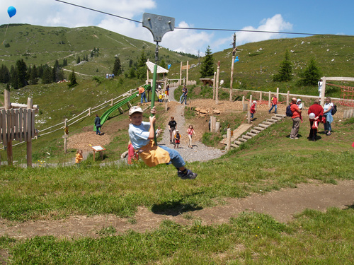 Ein Kind fährt auf einer Seilrutsche auf einem Spielplatz im Freien mit grasbewachsenen Hügeln und mehreren Personen im Hintergrund.