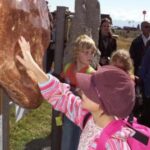 Ein Kind berührt einen großen, glatten Stein, der im Freien ausgestellt ist, während andere Leute im Hintergrund zusehen.