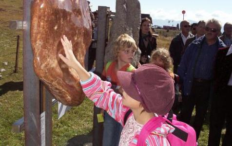 Ein Kind berührt einen großen, glatten Stein, der im Freien ausgestellt ist, während andere Leute im Hintergrund zusehen.