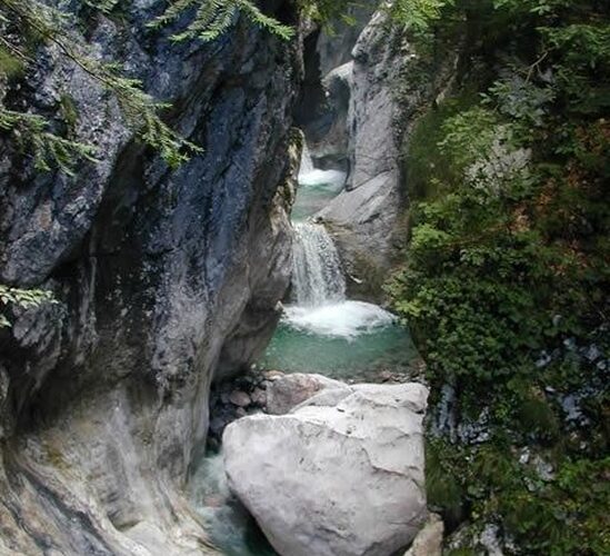 Ein kleiner Wasserfall fließt zwischen felsigen Klippen in einen klaren, türkisfarbenen Pool, der von üppiger grüner Vegetation umgeben ist.