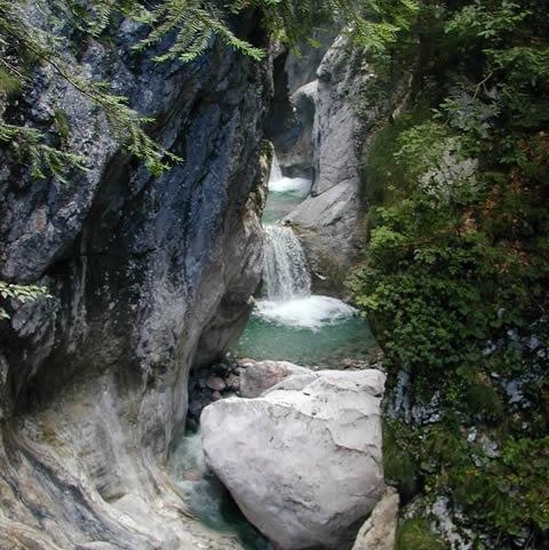 Ein kleiner Wasserfall fließt zwischen felsigen Klippen in einen klaren, türkisfarbenen Pool, der von üppiger grüner Vegetation umgeben ist.
