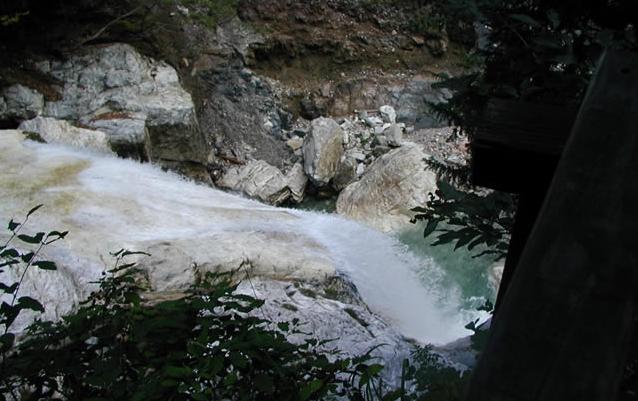 Ein kleiner Wasserfall fließt über weiße Felsen, die von Grün umgeben und teilweise von Bäumen beschattet sind.