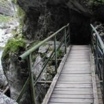 Eine schmale Holzbrücke mit Metallgeländer führt über eine felsige Schlucht, durch die ein Bach fließt.