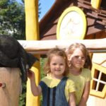 Zwei junge Mädchen stehen vor einem hölzernen Spielhaus; eines streichelt eine schwarze Katzenstatue auf einem Pfosten.