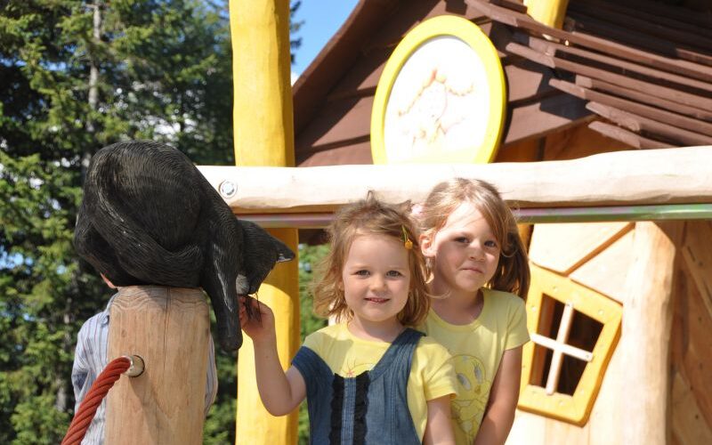 Zwei junge Mädchen stehen vor einem hölzernen Spielhaus; eines streichelt eine schwarze Katzenstatue auf einem Pfosten.
