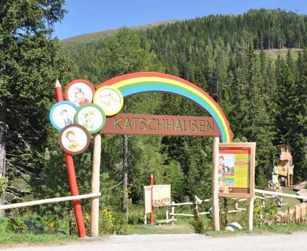 Eingang zum Erlebnispark Katschhausen mit einem Holzschild mit einem Regenbogen und Cartoon-Tieren, umgeben von Bäumen und einer Waldlandschaft.