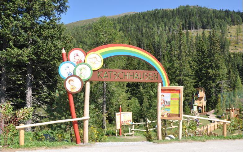 Eingang zum Erlebnispark Katschhausen mit einem Holzschild mit einem Regenbogen und Cartoon-Tieren, umgeben von Bäumen und einer Waldlandschaft.