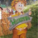 Eine Zeichentrickfigur mit einem Schild mit der Aufschrift "Willkommen" und einem Kaninchen, das hinter der Figur hervorlugt, steht im Freien auf einer Wiese mit einem Zaun und Bäumen im Hintergrund.